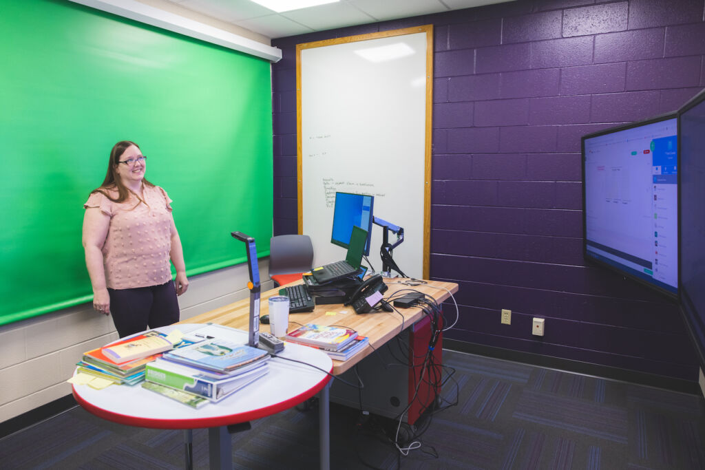In an instructional studio, a teacher stands behind her desk with a green screen in the background. She has a document camera and large monitor in front of her.