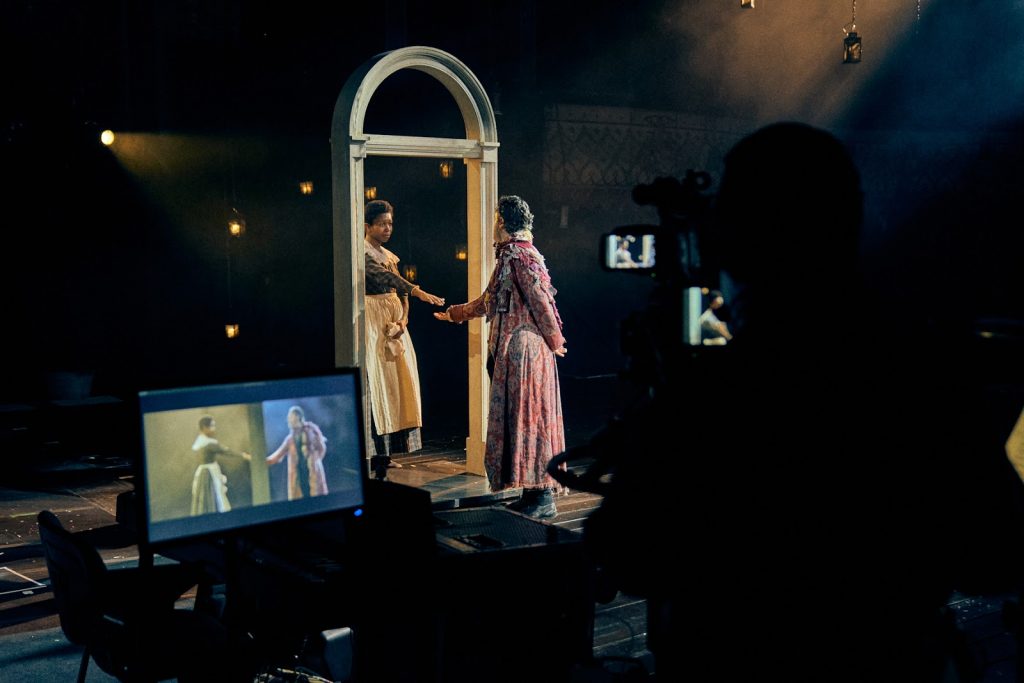 Dos actores en el escenario pasando por el arco de una puerta, con una pantalla de equipo que muestra a los actores en primer plano.